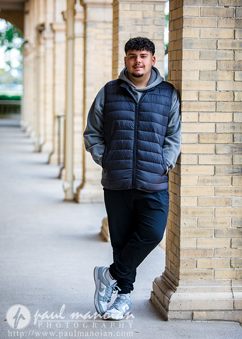 A young man with short curly hair leans casually against a brick column in a covered outdoor walkway during his Detroit senior portraits session, wearing a gray hoodie, black vest, black pants, and gray sneakers amid repeating columns.