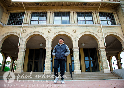 A man wearing a puffer jacket stands on the steps of a historic brick building with arched doorways and tall windows during a Detroit senior portraits session. The overcast sky highlights the structure’s classical architectural details.