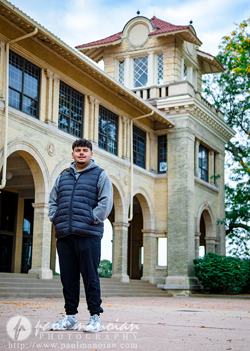 A young man wearing a dark vest, gray hoodie, and black pants stands confidently in front of a historic brick building with arches and a tall clock tower on a cloudy Detroit senior portraits session.