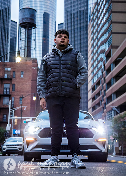 A young man in a gray hoodie and black vest stands confidently in front of a white sports car on a city street, surrounded by tall modern buildings under an overcast sky during his Detroit senior portraits session.