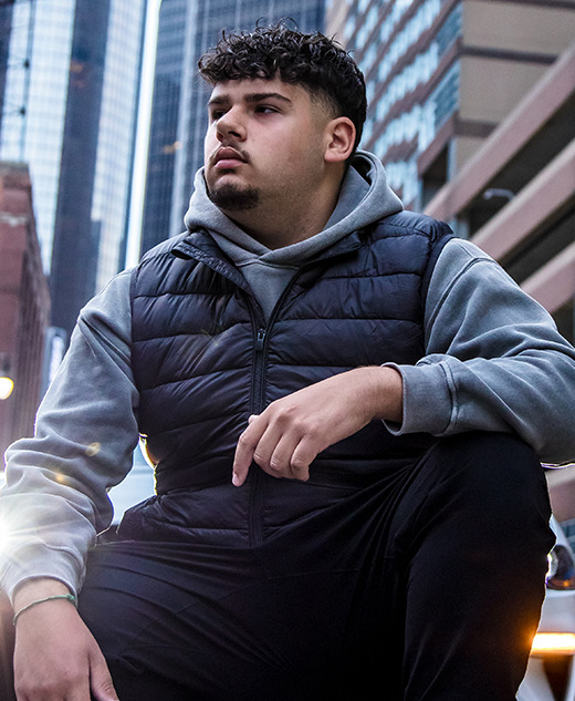 A young man with curly hair, wearing a gray hoodie and black vest, sits on a city street. Tall buildings and a Mustang with headlights on set the scene for this thoughtful Urban Session—perfect for Detroit Senior Pictures.