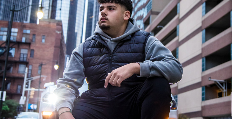 A young man with curly hair, wearing a gray hoodie and black vest, sits on a city street. Tall buildings and a Mustang with headlights on set the scene for this thoughtful Urban Session—perfect for Detroit Senior Pictures.