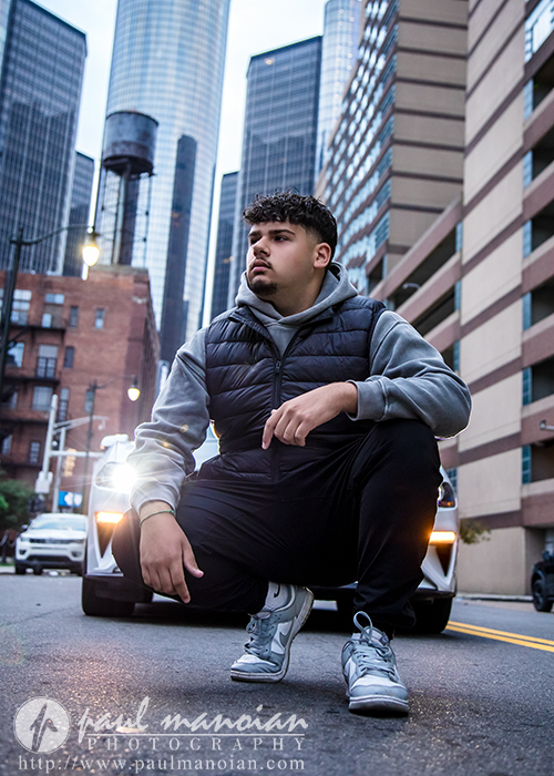 A young man squats confidently in the middle of a city street during his Detroit senior portraits session, with tall skyscrapers and a parked car behind him. He wears a gray hoodie, black vest, and sneakers. Urban buildings surround the scene at dusk.