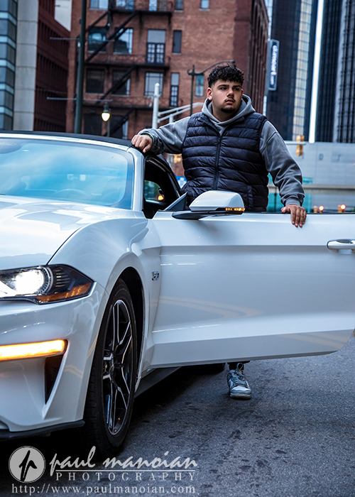 A young man in a gray hoodie and black vest stands next to an open white car door on a city street, with tall buildings in the background, captured during a Detroit senior portraits session. A photography watermark appears at the bottom of the image.
