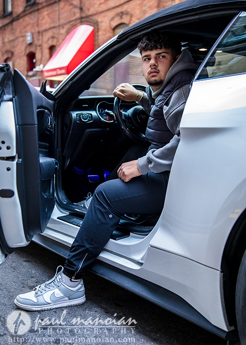A young man sits in the driver’s seat of a white car with the door open, looking to his left during his Detroit senior portraits session. He wears a grey jacket, dark pants, and Nike sneakers against a brick building backdrop.