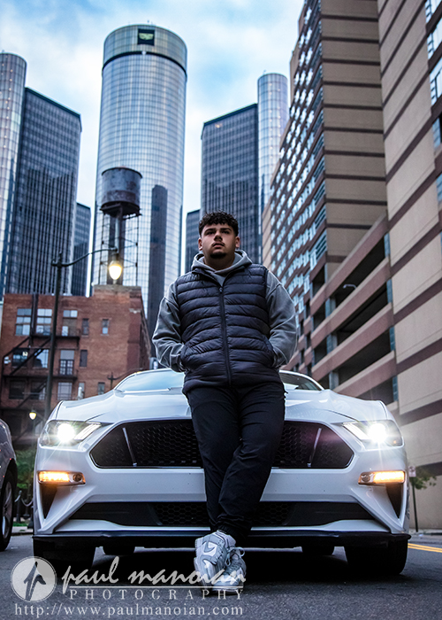 A young man in a dark vest and hoodie leans against the front of a white sports car parked on a city street, skyscrapers rising behind him—capturing the essence of a Detroit senior portraits session.