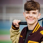 A smiling teenage boy in a letterman jacket holds a tennis racket over his shoulder, standing on a tennis court during his tennis senior pictures session.
