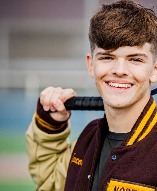 A smiling teenage boy in a letterman jacket holds a tennis racket over his shoulder, standing on a tennis court during his tennis senior pictures session.