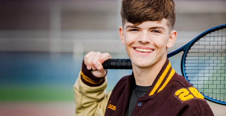 A smiling teenage boy in a letterman jacket holds a tennis racket over his shoulder, standing on a tennis court during his tennis senior pictures session.