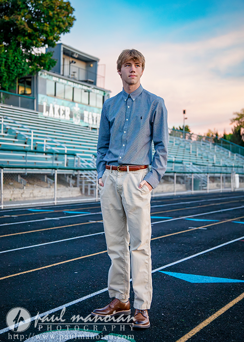 A young man in a blue button-down shirt and beige pants stands on a running track near empty bleachers, with a sunset sky in the background.