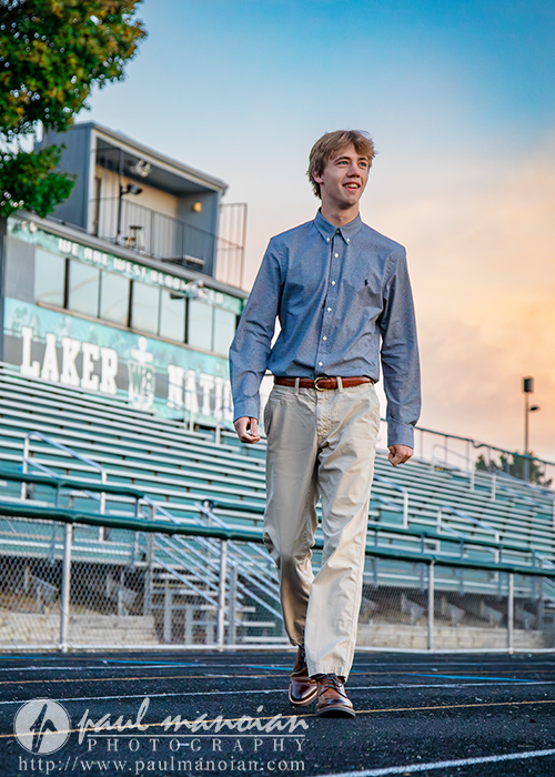 A young man in a blue shirt and beige pants walks confidently on a track in front of empty bleachers at a stadium during sunset.