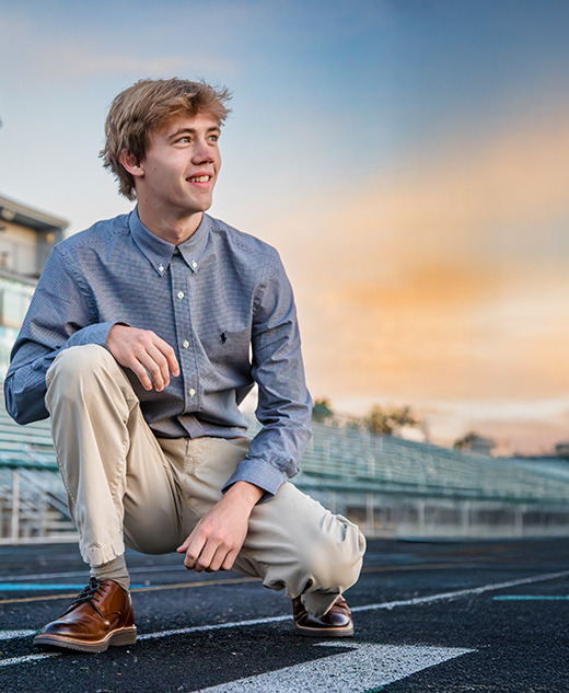 Sebastien kneels on a track in a blue button-up shirt and beige pants, smiling as he gazes to the side. The empty bleachers and sunrise sky create a vibrant backdrop, perfect for West Bloomfield Senior Pictures.