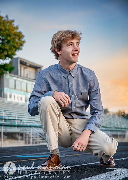 A young man in a blue button-down shirt and khaki pants crouches on a track field, smiling and looking to the side. Bleachers and a building are in the background under a colorful sunset sky.