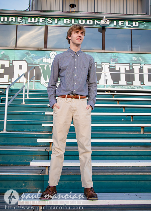 A young man in a blue button-down shirt and beige pants stands on bleachers in front of a sports field sign that reads "WEST BLOOMFIELD FIELD." He looks to the side and smiles.