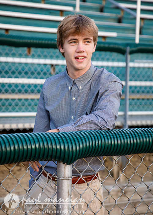 A young man with light brown hair, wearing a button-down shirt and khaki pants, stands and leans against a green and silver chain-link fence in an outdoor stadium. Empty bleachers are visible in the background.
