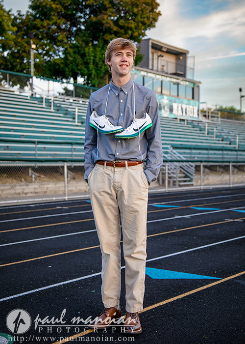 A young man stands on a running track, smiling, with running shoes draped around his neck. He wears a blue button-down shirt and khaki pants. Bleachers and a press box are visible in the background.