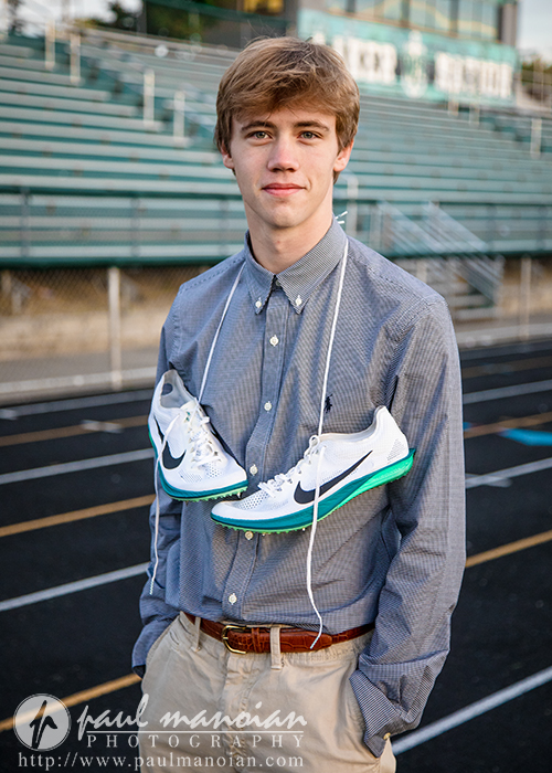 A young man standing on a running track, wearing a button-up shirt and khakis, with a pair of white Nike running shoes hanging around his neck. Bleachers are visible in the background.