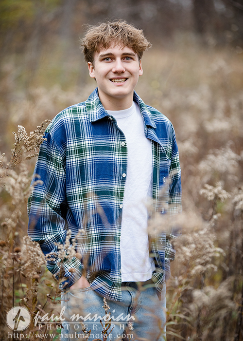 A young man with short, wavy hair smiles while standing in a field of tall, dry grass during his Ann Arbor senior pictures session. He wears a blue and green plaid shirt over a white T-shirt and jeans. The background is blurred and natural.