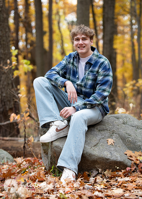 A young man in a blue plaid shirt and light jeans sits smiling on a large rock in a forest filled with autumn leaves and golden foliage during his Ann Arbor senior pictures session.