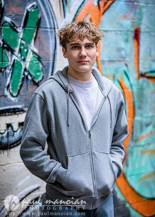 A young person with short, curly hair wearing a gray zip-up hoodie and white t-shirt stands in front of a colorful graffiti-covered wall, looking at the camera with hands in pockets during an Ann Arbor senior pictures session.