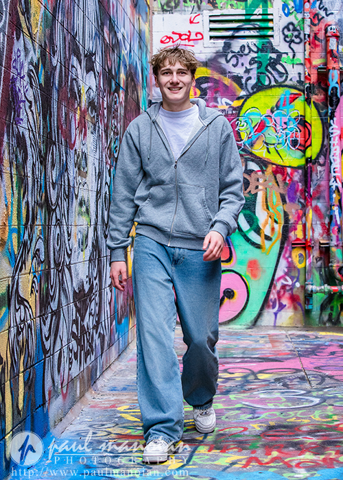 A young man in a gray hoodie and blue jeans smiles as he walks down a narrow alley covered in colorful graffiti art during his Ann Arbor senior pictures session.