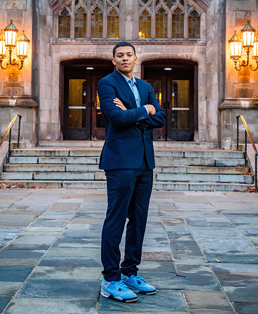 A man in a suit stands confidently with arms crossed on a stone walkway during his Greenhills senior pictures session, in front of an ornate building entrance illuminated by warm lanterns.