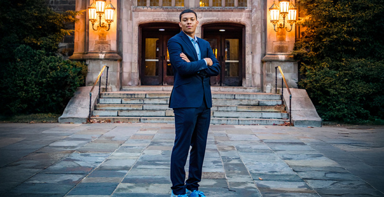 A man in a suit stands confidently with arms crossed on a stone walkway during his Greenhills senior pictures session, in front of an ornate building entrance illuminated by warm lanterns.