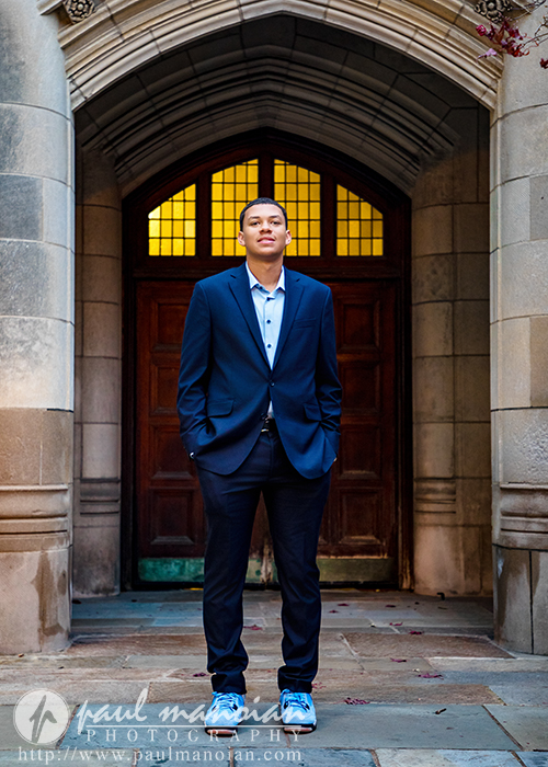 A high school senior in a blue suit and blue sneakers stands confidently in front of a large wooden door with arched stone architecture and yellow stained glass windows during his Greenhills senior pictures session.