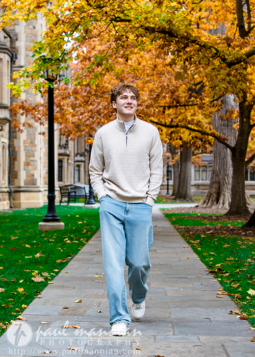 A young man in a light sweater and jeans walks on a stone path through a campus with autumn trees and historic buildings, smiling and looking upward during his Ann Arbor senior pictures session.