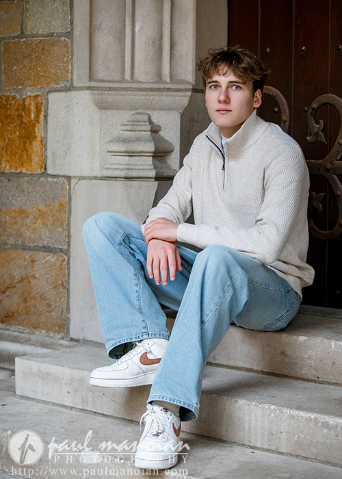 A young man in a light sweater and blue jeans sits on stone steps by a wooden door during his Ann Arbor senior pictures session, looking slightly to the side. He has short brown hair and wears white sneakers.