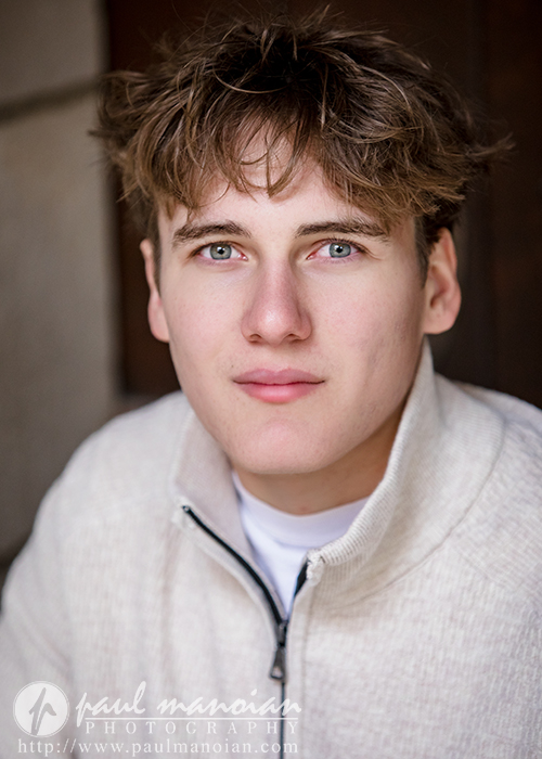 A young man with light brown, tousled hair and blue eyes wears a light beige zip-up sweater, looking directly at the camera with a neutral expression during his Ann Arbor senior pictures session. The softly blurred background and photography watermark are visible.