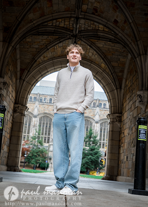 A young man wearing a beige sweater and light blue jeans stands under a stone archway during his Ann Arbor senior pictures session, with a historic building and trees in the background. Two caution signs are posted on pillars nearby.
