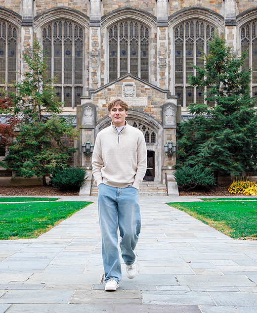 A young man in a light sweater and jeans walks along a stone pathway on a college campus during his Ann Arbor senior pictures session, with a large, historic building and colorful autumn trees in the background.