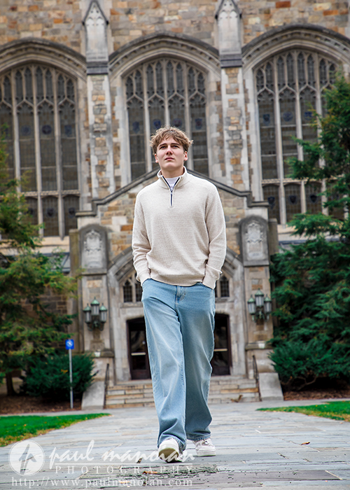 A young man in a cream sweater and blue jeans walks on a stone path in front of a large stone building with arched windows and a wooden door, surrounded by greenery during his Ann Arbor senior pictures session.