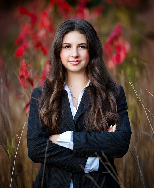 A high school senior with long brown hair, wearing a black suit and white shirt, stands confidently with arms crossed in front of tall grass and red autumn foliage during her Ann Arbor senior photos session.
