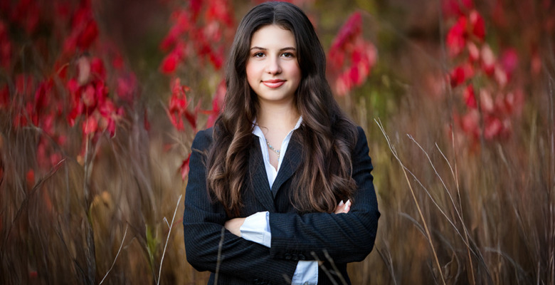 A high school senior with long brown hair, wearing a black suit and white shirt, stands confidently with arms crossed in front of tall grass and red autumn foliage during her Ann Arbor senior photos session.