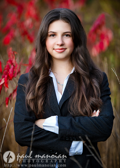A young woman with long brown hair, wearing a black pinstripe suit and white shirt, stands outdoors with her arms crossed, smiling slightly during her Ann Arbor senior photos session. Red and green foliage is blurred in the background.