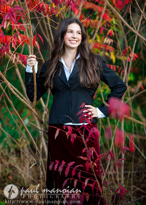 A high school senior with long brown hair, wearing a black blazer and red skirt, stands outdoors among vibrant red and green autumn leaves during her Ann Arbor senior photos session, smiling and holding a thin tree branch.