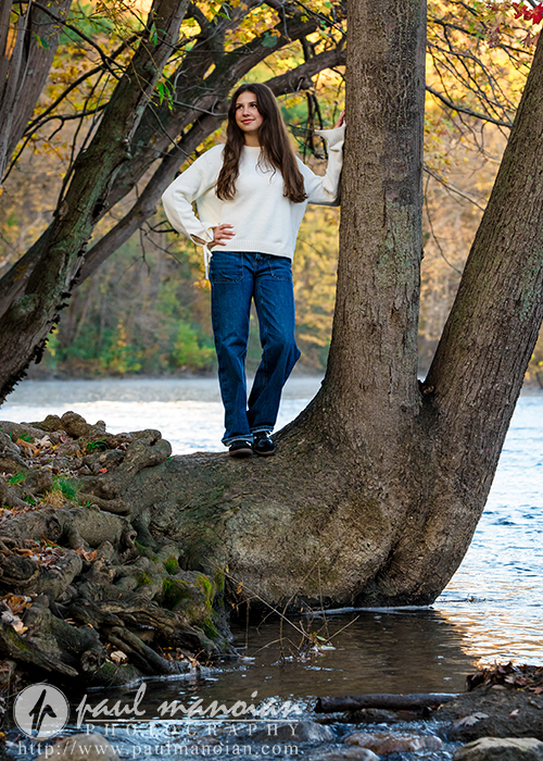 A high school senior in a white sweater and blue jeans stands on the roots of a tree by a river during her Ann Arbor senior photos session, with autumn leaves and water in the background. She rests one hand on the trunk and smiles into the distance.