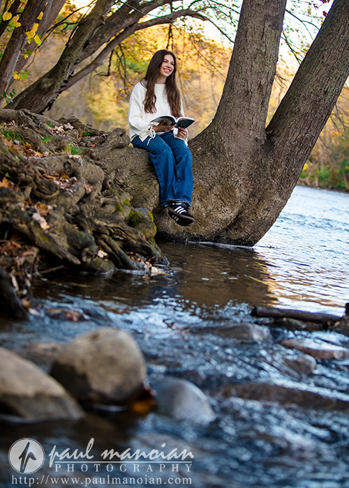 A high school senior sits on a large tree trunk beside a river, smiling while reading a book during her Ann Arbor senior photos session. She wears a white sweater and blue jeans, surrounded by rocks, trees, and water on a sunny day.