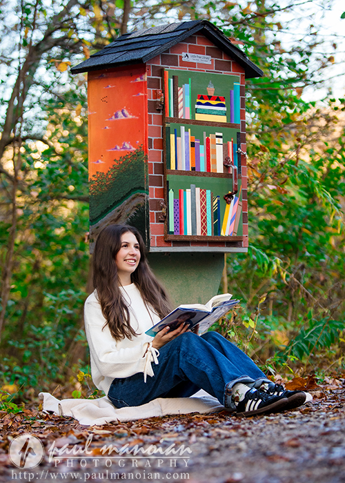 A high school senior sits on a blanket outdoors, reading beside a colorful little free library decorated with painted books and a sunset—an ideal Ann Arbor senior photos session scene, surrounded by lush green foliage and trees.