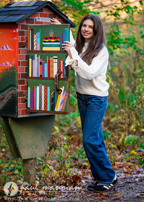 A high school senior in a white sweater and jeans stands outdoors, smiling and holding open the door of a small outdoor library decorated with colorful book illustrations on a wooded path during her Ann Arbor senior photos session.