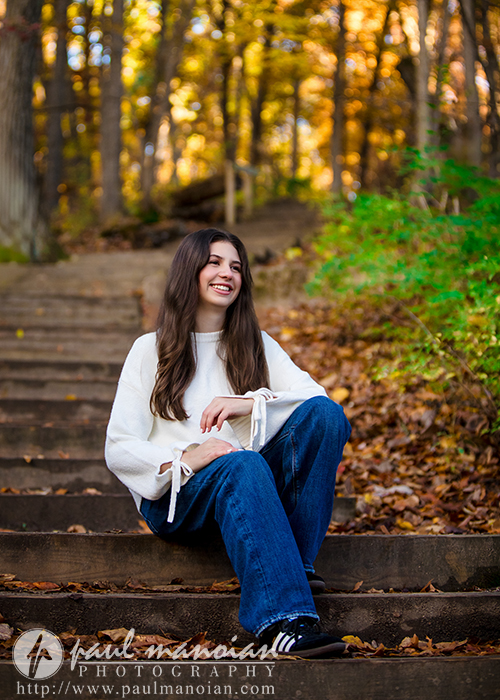 A high school senior with long brown hair, wearing a white sweater and blue jeans, sits on outdoor stone steps surrounded by autumn trees and fallen leaves, smiling and looking to the side during her Ann Arbor senior photos session.