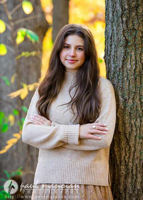 A high school senior with long brown hair, wearing a beige sweater, stands outdoors leaning against a tree with arms crossed during her Ann Arbor senior photos session. Autumn leaves and sunlight brighten the background.