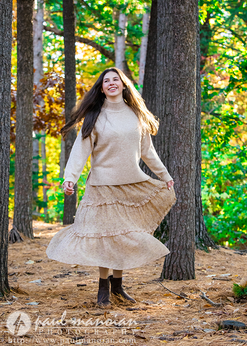 A high school senior in a beige sweater and long skirt twirls joyfully among tall pine trees in a sunlit forest during an Ann Arbor senior photos session, with fallen needles carpeting the ground and green foliage in the background.