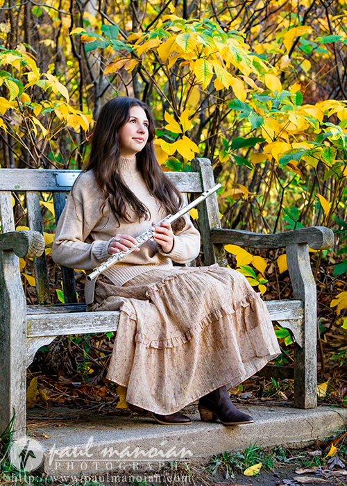 A high school senior with long brown hair sits on a wooden bench outdoors, holding a flute during her Ann Arbor senior photos session. She wears a beige sweater and a long tan skirt, surrounded by yellow autumn leaves and greenery in the background.
