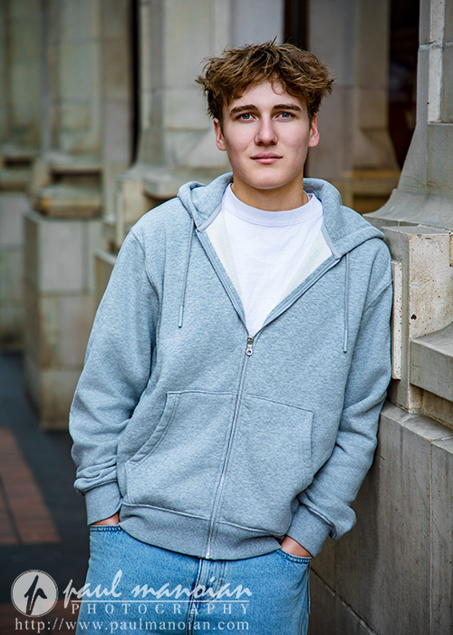 A young man with curly brown hair wearing a gray hoodie and white t-shirt leans against a stone building, looking at the camera with a relaxed expression during his Ann Arbor senior pictures session. The logo "paul manarian photography" appears at the bottom.