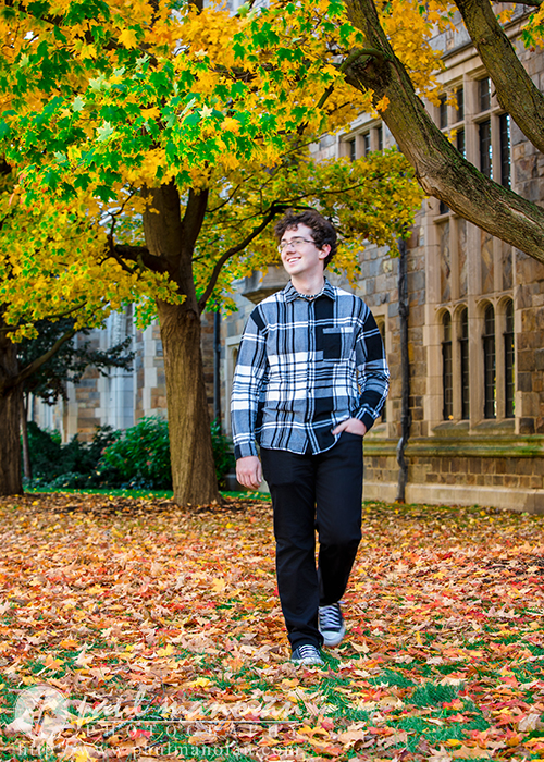 A young man wearing a black-and-white plaid shirt and black pants walks through fallen autumn leaves under yellow-leaved trees, with a stone building in the background during his South Lyon senior pictures session.