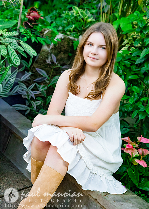 A high school senior with long, light brown hair sits on a stone edge in a lush garden during her Grosse Pointe senior pictures session, wearing a sleeveless white dress and tan boots. She smiles softly, surrounded by green foliage and pink flowers.