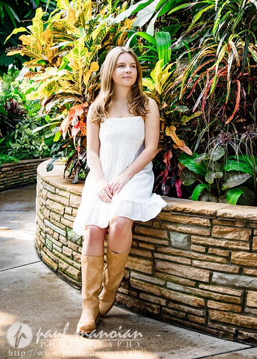 A high school senior in a white dress and tan boots sits on a stone bench surrounded by lush green and yellow tropical plants, looking off to the side with a peaceful expression during her Grosse Pointe senior pictures session.